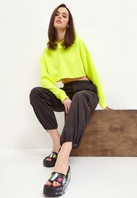 Hanorac scurt de culoare verde neon, pantaloni negri largi cu manșete elastice și sandale negre masive cu accente grafice colorate.