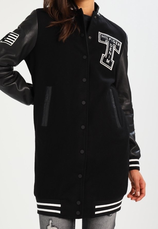 Kobieta ubrana w długą czarną kurtkę varsity z skórzanymi rękawami, naszywkami na piersi i rękawie oraz prążkowanymi mankietami i dolną krawędzią.