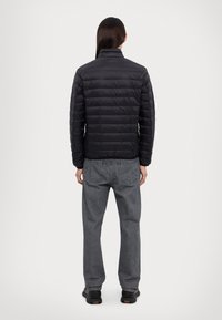 Veste rembourrée noire avec un col haut, présentant une texture matelassée, portée sur un jean gris. Vue de dos. Aucun matériel ou motif visible.