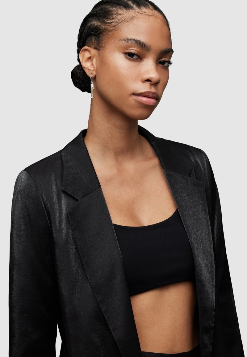Schwarze Satin-Blazer mit strukturiertem Design und retournierten Revers. Über einem schwarzen Cropped-Top getragen, das eine klare, moderne Silhouette betont.
