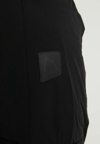 Schwarzer Stoff mit einem kleinen schwarzen quadratischen Aufnäher, der ein geprägtes minimalistisches Logo zeigt und auf der Oberfläche aufgenäht ist.