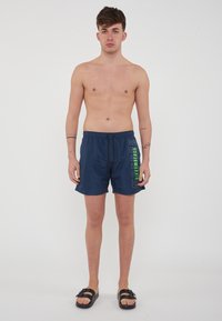 Marineblauwe zwembroek van lichtgewicht stof, met een trekkoord taille en een opvallend groen tekstontwerp aan de voorkant.