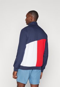 Navy zip-up jacket with a color block design featuring red and white accents, made of smooth fabric, with a high collar and ribbed cuffs.
