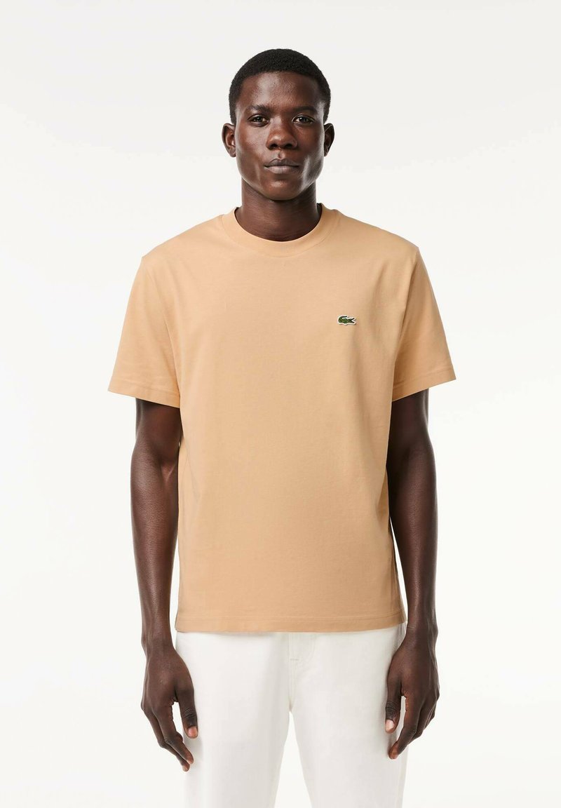 T-shirt in cotone beige con maniche corte e scollo rotondo. Presenta un piccolo logo verde sul petto. Texture liscia, design a vestibilità standard.