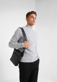 Maglione grigio chiaro con collo alto a costine, abbinato a pantaloni neri e una borsa a tracolla in pelle nera con tasca con zip.