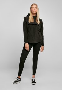 Pullover in pile nero con cappuccio, zip a metà e tasca frontale, abbinato a leggings neri e sneakers nere con suola bianca.