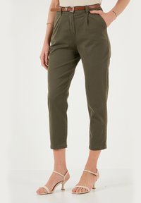 Pantaloni verde oliva a vita alta e a cropped, dotati di una cintura marrone, in morbido tessuto di cotone e con dettaglio plissettato davanti. Abbinati a tacchi beige.
