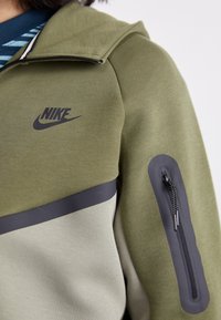 Olívazöld kapucnis pulóver fekete Nike logóval, sötétszürke csíkkal és cipzáras zsebbel a felkaron. Simas�ágra és kellemes tapintású anyag.