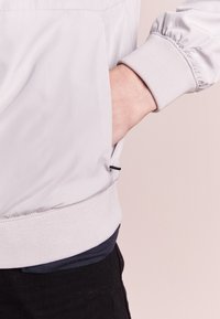 Chaqueta gris clara con textura suave, puños elásticos y dobladillo acanalado; mano en el bolsillo lateral, superpuesta sobre una camisa oscura.