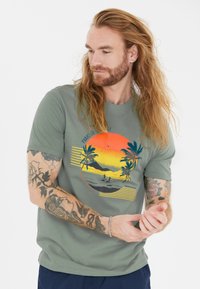 Vihreä puuvillapaita, jossa on auringonlaskugrafiikka palmupuineen, raitoja ja rannikkoskenaario. Lyhyet hihat ja rento istuvuus.