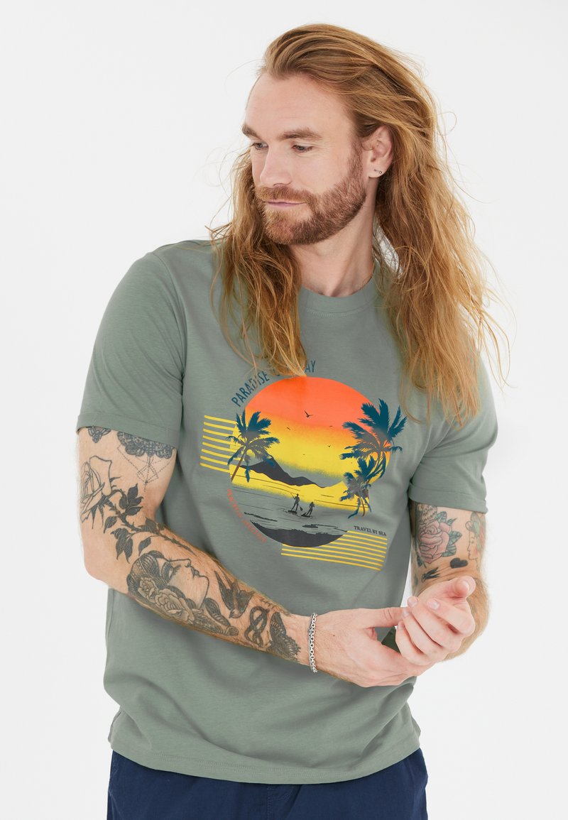 Vihreä puuvillapaita, jossa on auringonlaskugrafiikka palmupuineen, raitoja ja rannikkoskenaario. Lyhyet hihat ja rento istuvuus.