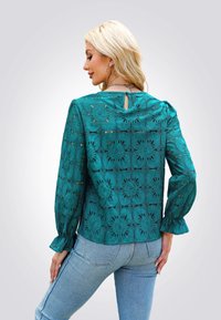 Blusa teal con un design in pizzo floreale, maniche lunghe a sbuffo e dettaglio a occhiello sul retro, abbinata a jeans azzurri.