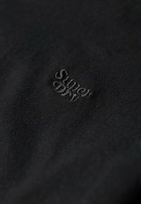 Tessuto di cotone nero con un logo ricamato "Superdry" in filo scuro, cucito su una superficie liscia senza ulteriori motivi.