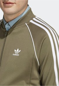 Oliwkowo-zielona sportowa kurtka z białymi paskami, zapinana na zamek, z prążkowanym kołnierzem i haftowanym logo Adidas na lewej piersi. Gładka faktura.