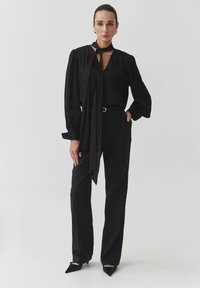 Black blouse with long sleeves and a tie neckline, paired with high-waisted black trousers. The outfit features a sleek, tailored design.