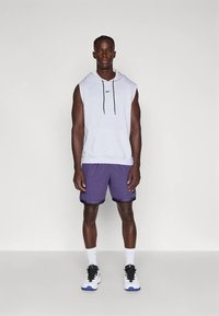 Γκρίζα αμάνικη ζακέτα με μαύρα κορδόνια, μοβ τρυπητά σορτς, λευκές κάλτσες και μαύρα-λευκά αθλητικά παπούτσια με μπλε λεπτομέρειες.