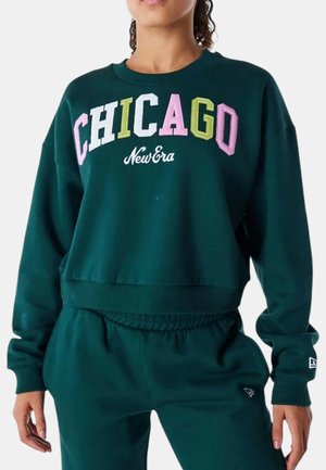 Donna che indossa felpa verde scuro e pantaloni, la felpa ha "CHICAGO" in lettere multicolore e "New Era" in scrittura bianca.