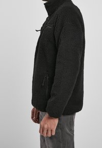 Schwarze Fleecejacke mit hohem Kragen, Reißverschluss-Brusttasche, Seitentaschen und strukturiertem Stoff. Mit verstellbaren Kordeln am Saum.
