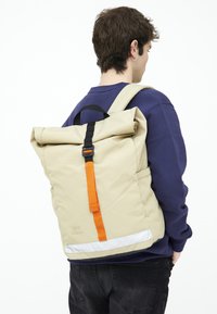 Beige rulltop ryggsäck med orange rem och svarta detaljer. Har en texturerad yta, justerbara remmar och reflektionsdetaljer.