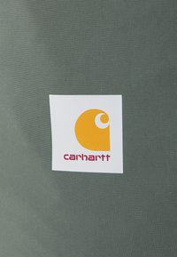 Zelena tkanina u pozadini s bijelom kvadratnom oznakom na kojoj je stilizirano žuto slovo "C" i crveni tekst "carhartt" na dnu.