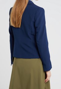 Navy blue fitted blazer with structured shoulders and long sleeves, paired with a soft olive green skirt. Smooth fabric with visible seams.