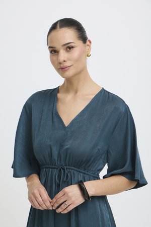 Donna con capelli scuri raccolti indossa orecchini d'oro, un vestito color verde acqua con scollo a V e maniche svolazzanti, un braccialetto nero e un anello alla mano sinistra.