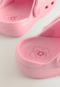 Roosad plastmassist sandaalid tekstuurse tallaga, lillekujuline disain sisetallal ja reguleeritav rihm. Märgatav täpiline pind ja siledad servad.