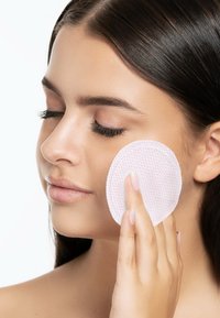 Tampon en coton avec des points roses tenu contre la joue d'un modèle. Peau lisse, maquillage naturel et cheveux noirs raides sont visibles.