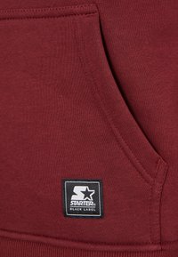 Gros plan d'une poche en tissu grenat avec des coutures visibles et un patch noir du logo Starter Black Label cousu dessus.