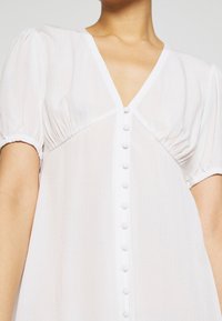 Vestido blanco de mangas abullonadas con escote en V, tejido texturizado y una fila de botones decorativos en la parte delantera.