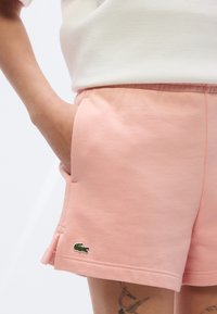Shorts en coton rose avec taille élastique, poches latérales et logo vert brodé près de l'ourlet. Texture lisse, design décontracté.
