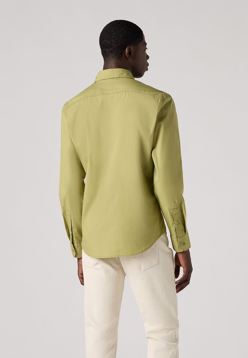 Chemise à manches longues, vert olive clair, avec un col pointu, un ourlet droit et des poignets boutonnés, associée à un pantalon couleur crème.