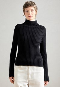 Pull à col roulé noir à côtes avec des manches longues, présentant une silhouette ajustée et un col montant, associé à un pantalon de couleur claire.