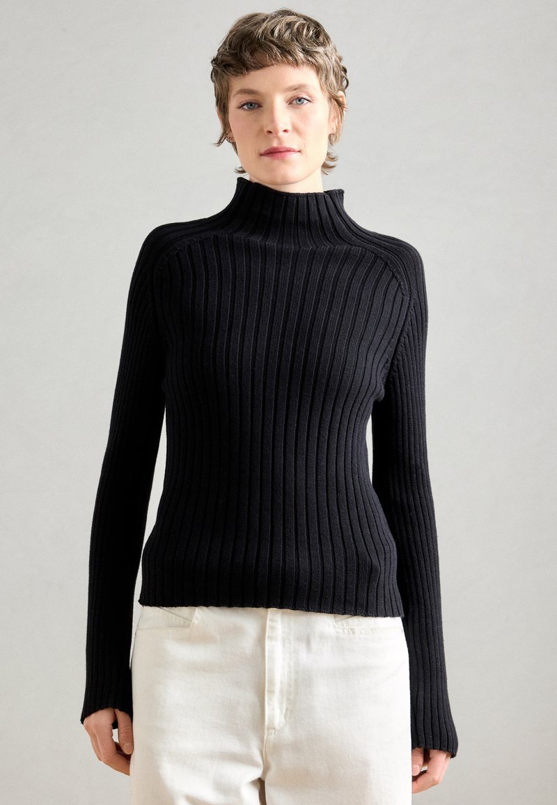 Pull à col roulé noir à côtes avec des manches longues, présentant une silhouette ajustée et un col montant, associé à un pantalon de couleur claire.