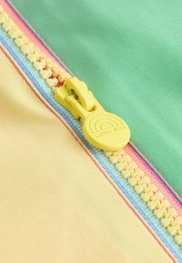 Rumeno jeziček zadrge z napisom »Little Bird« na pastelno rumenem, zelenem in rožnatem blagu, prikazujoč podrobne zobce zadrge in šive.