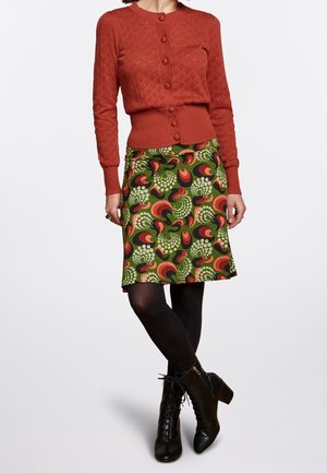 Femme portant un cardigan boutonné rouge rouille, une jupe à motifs verts et rouges, des collants noirs et des bottines à lacets noires.