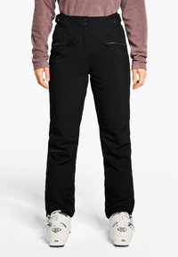 Person wearing black high-waist ski pants with zippered pockets, a mauve long-sleeve top, and white ski boots on a white background.