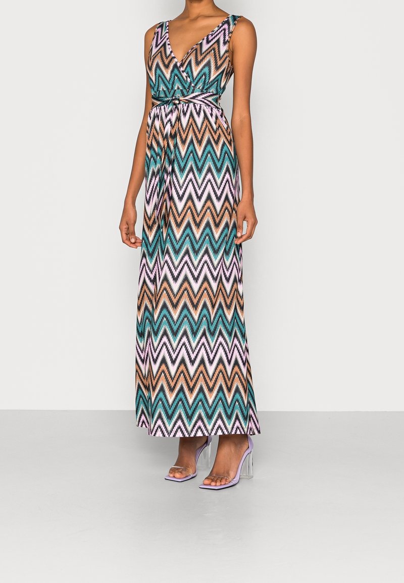 Femme portant une robe maxi sans manches en V avec des motifs zigzag colorés en bleu sarcelle, marron, blanc et violet, associée à des chaussures à talons transparents.