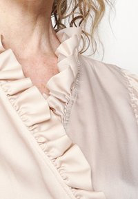 Blouse beige clair avec un col volanté, présentant une texture de tissu lisse. La vue rapprochée montre le détail du design des volants et des coutures.