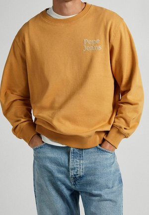 Person trägt einen senffarbenen Pepe Jeans Pullover und hellblaue Jeans, die Hände in den Taschen, vor einem schlichten Hintergrund.