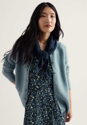 Femme aux longs cheveux noirs portant une robe bleue fleurie, un cardigan bleu clair boutonné et une écharpe bleu marine nouée autour du cou, debout devant un fond uni.