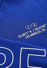 Blå sweatshirt med vit text som visar koordinater, "33.9871° N / 118.4755° W," och "LOS ANGELES, CA" nära fållen.