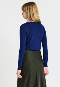 Frau mit blonden Haaren, die ein blaues Langarmshirt und einen hoch-taillierten dunkelgrünen Rock trägt, blickt nach links vor einem weißen Hintergrund.