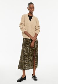 Vaaleanbeige neulekangastakki, jossa on ribbitekstuuri ja kaksi nappia, yhdistetty mustaan kukkakuvioiseen maksihameeseen, jossa on sivuaukko. Mustat loafersit viimeistelevät asun.