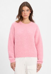 Jonge vrouw met blauwe ogen, gekleed in een gestructureerde roze trui en witte broeken met een grote zijzak, staande tegen een effen achtergrond.
