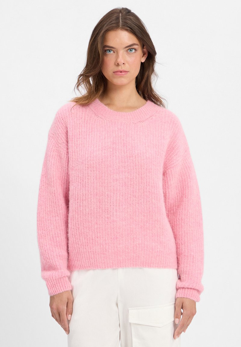 Jonge vrouw met blauwe ogen, gekleed in een gestructureerde roze trui en witte broeken met een grote zijzak, staande tegen een effen achtergrond.