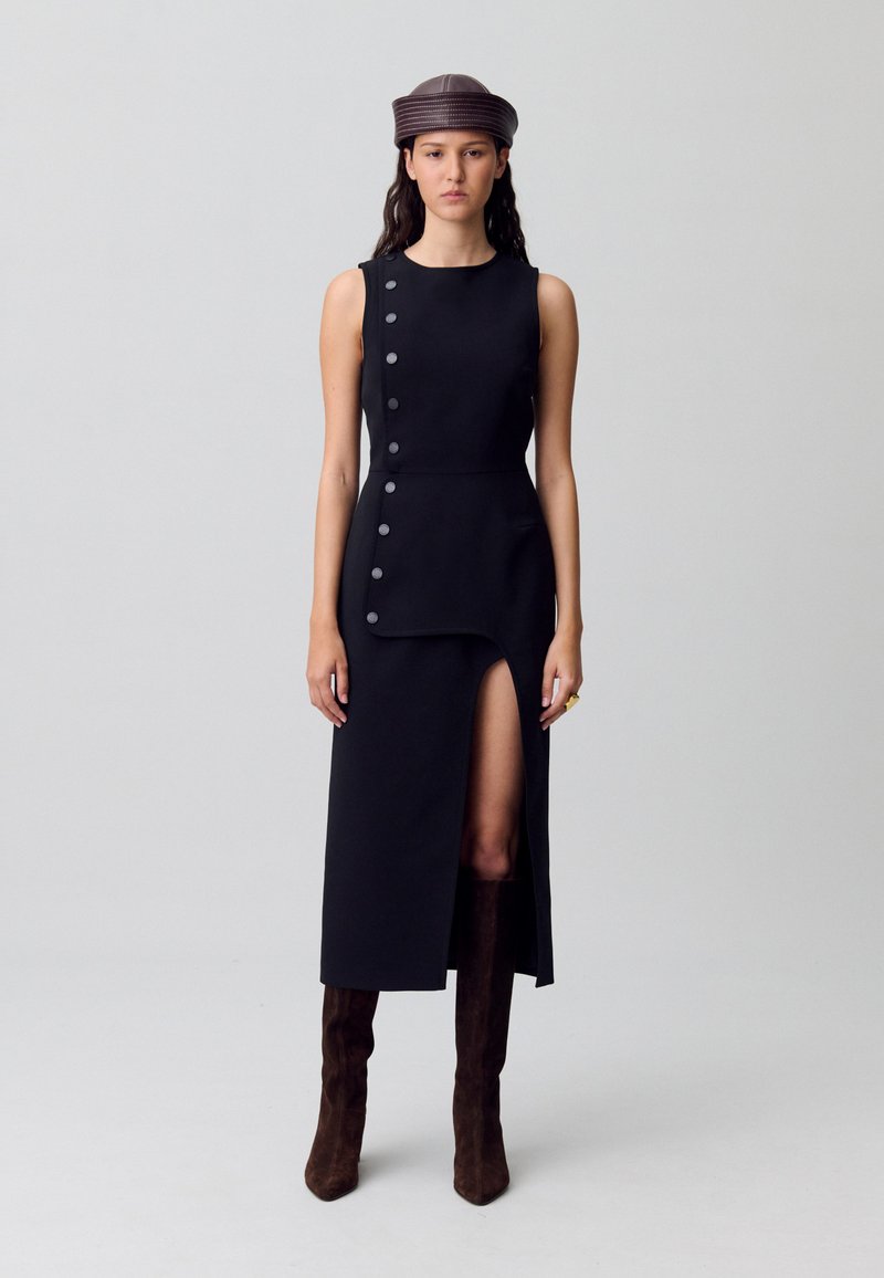 Vestido negro sin mangas con una abertura frontal, que presenta grandes botones en un lado. La modelo lleva un sombrero marrón a juego y botas altas de ante.