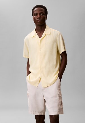 Mann trägt ein hellgelbes kurzärmeliges Hemd mit Knöpfen und beige Cargo-Shorts, steht mit Händen in den Taschen vor einem schlichten Hintergrund.