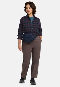 Camicia a quadri bordeaux e verde, borsa in vita blu navy, pantaloni beige con una texture liscia e scarpe sportive nere.