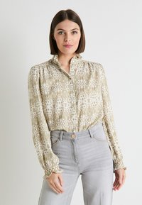 Camisa de botones en color beige claro con un patrón intrincado. Mangas largas con puños con volantes. Combinada con unos jeans de tiro alto en gris claro.